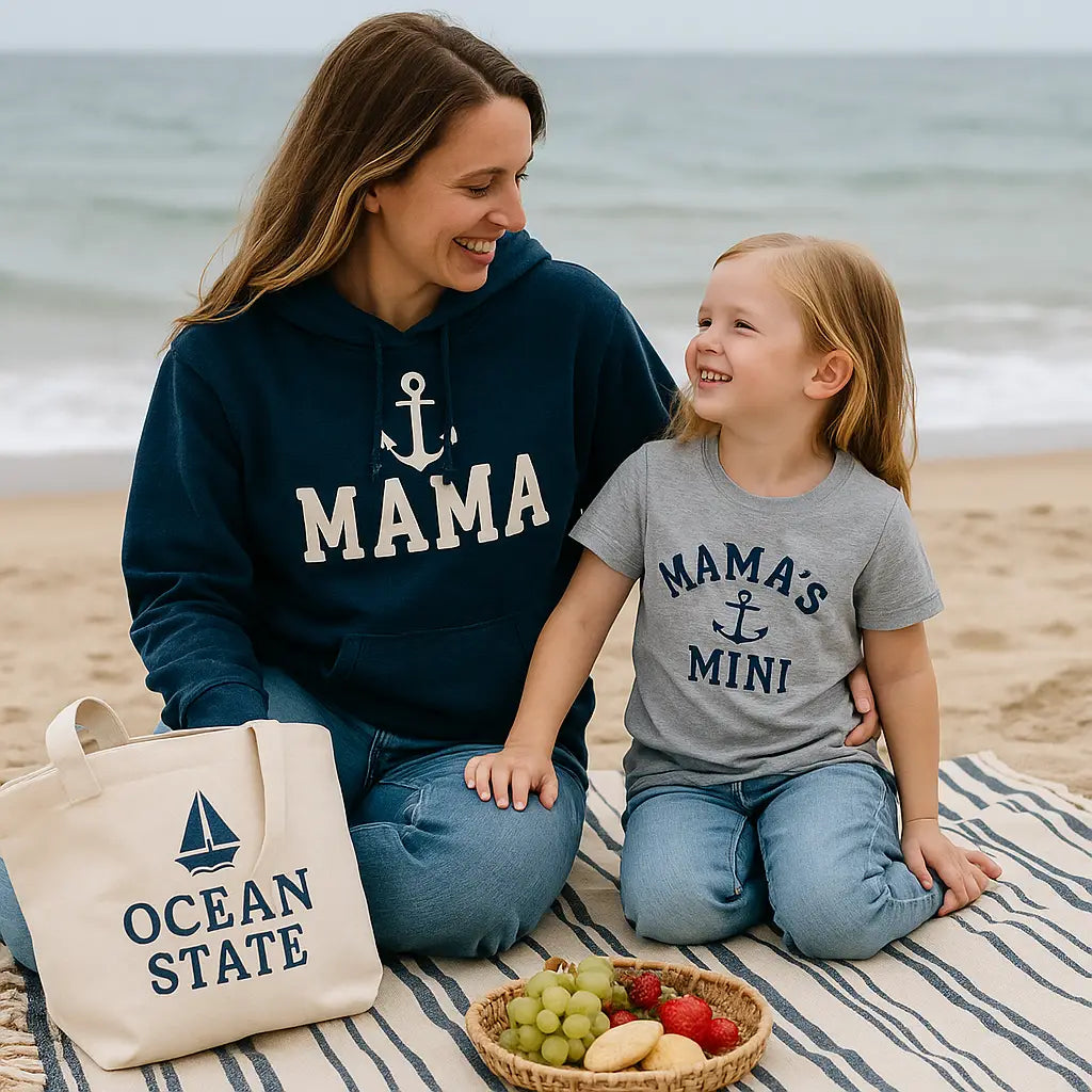 A realistic beach picnic scene. A mother and child wearing matching coastal-inspired apparel (nautical hoodie and tee), sitting on a blanket with tote bag nearby. Ocean visible in background, soft natural light, warm family aesthetic.
